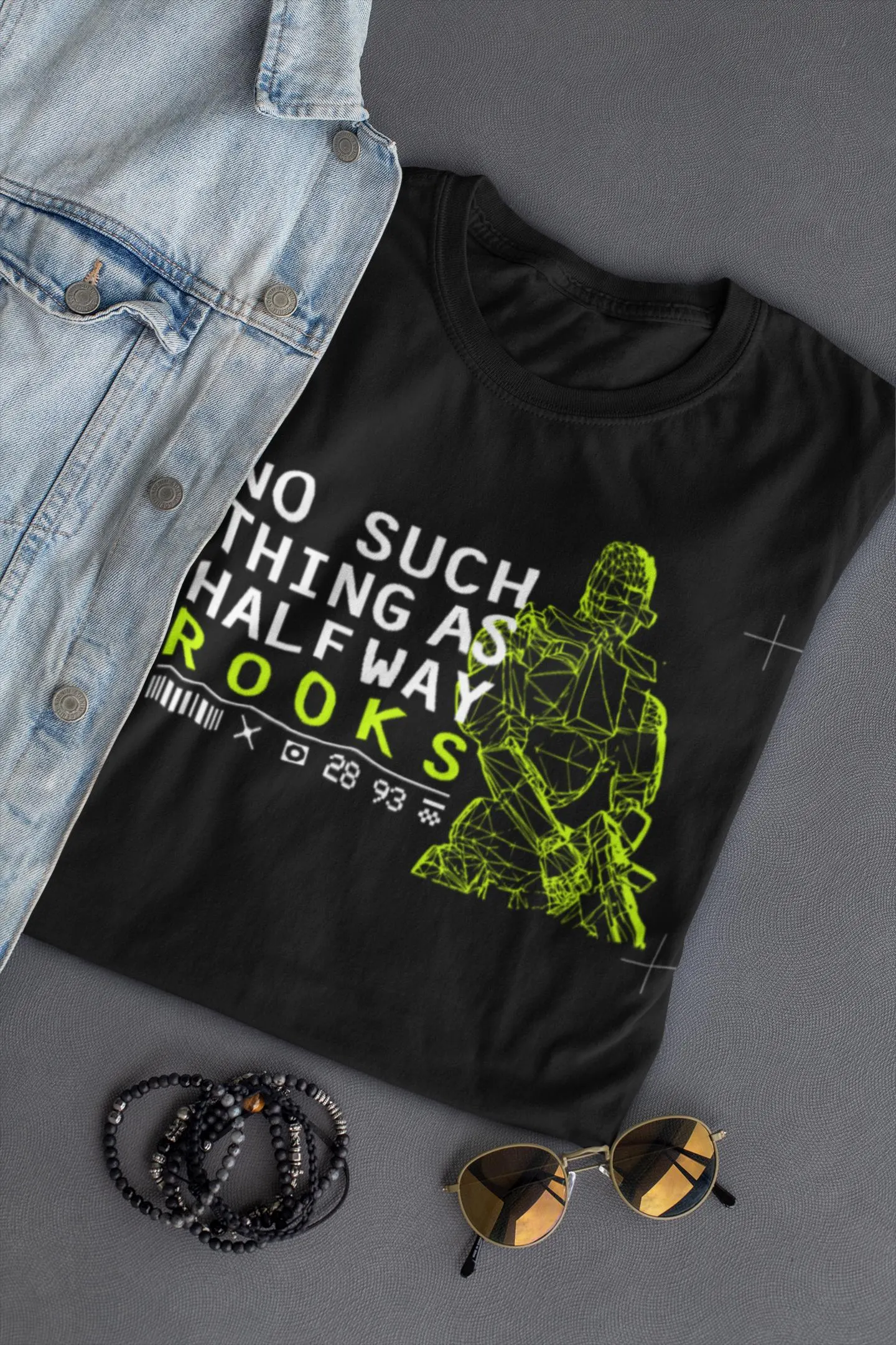 Black t-shirt with graphic design and text, placed on a gray surface with sunglasses and bracelet.