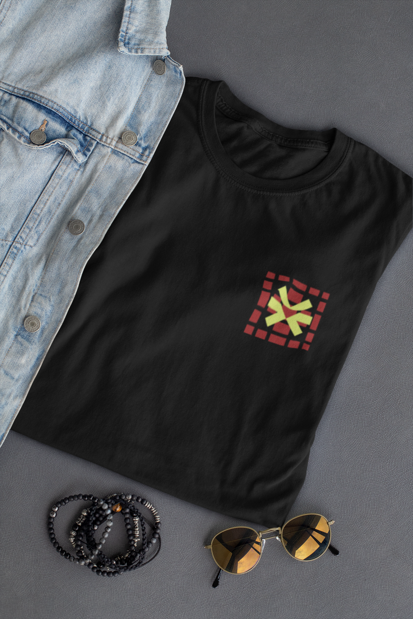 Black t-shirt with a colorful square design, denim jacket, sunglasses, and beads on a gray surface.